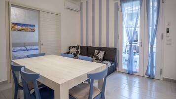 Una camera moderna con un tavolo da pranzo in legno, sedie blu, un divano con cuscini decorati e una porta scorrevole in vetro che dà su una zona esterna.