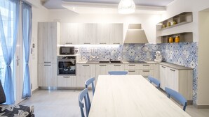 A modern kitchen with light wood cabinets, a white countertop, and blue and white patterned backsplash. There is a dining table with chairs, a hanging light fixture, and a window with curtains.