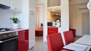 A modern kitchen with red cabinetry, a stove, and a microwave. There is a dining area with red chairs and a white table. - Apartment 100 meters from the beach (Gatteo A Mare)