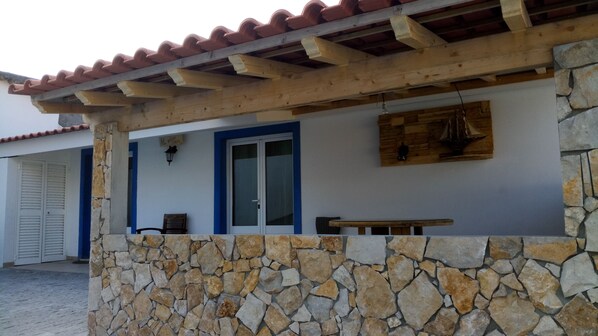 A stone wall with a wooden bench and a table, a wooden pergola with a tiled roof, and a door with a blue frame. - CASA DAMÉLIA POLO2-House near Óbidos in the slope of recycling décor (Vau Óbidos)