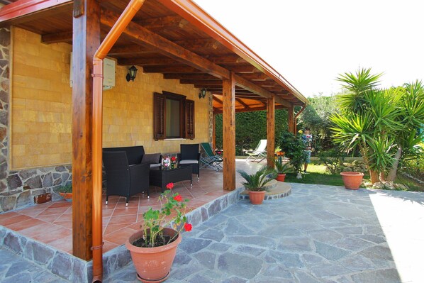 A covered outdoor seating area with wicker furniture, a stone floor, and potted plants.