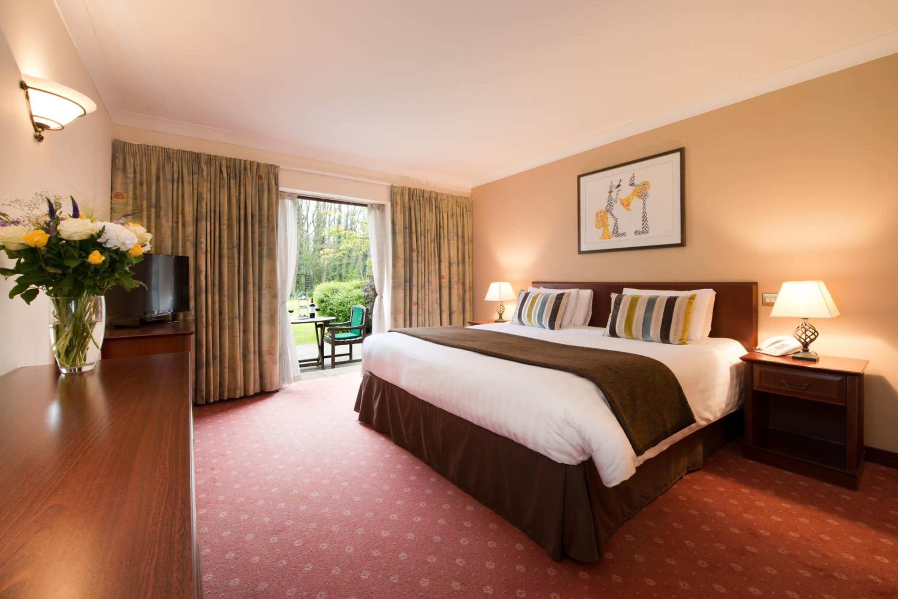 A hotel room with a bed, bedside tables, a desk with a TV, a vase with flowers, and a window with curtains.