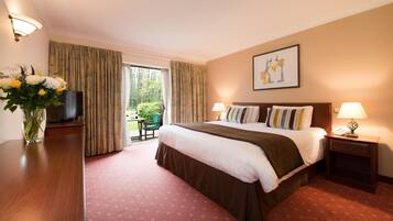 A hotel room with a bed, bedside tables, a desk with a TV, a vase with flowers, and a window with curtains.