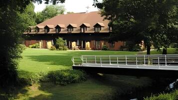 A building with a symmetrical roofline and multiple windows, surrounded by a well-maintained lawn and a small bridge over a water feature.