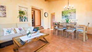 A living room with a sofa, coffee table, and dining table with chairs. There is a clock on the wall and a plant on the coffee table. - VILVAL Finca for 4 with pool in Moscari (Moscari)