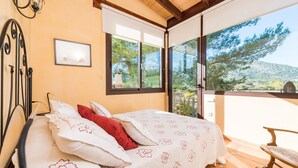 A bedroom with a bed, a nightstand, a window with a view of trees and hills, and a balcony with a chair. - VILVAL Finca for 4 with pool in Moscari (Moscari)