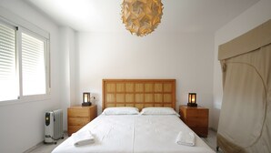 Un dormitorio ordenado con cabecera de madera, dos mesitas de noche con lámparas, un radiador y una ventana con persianas.