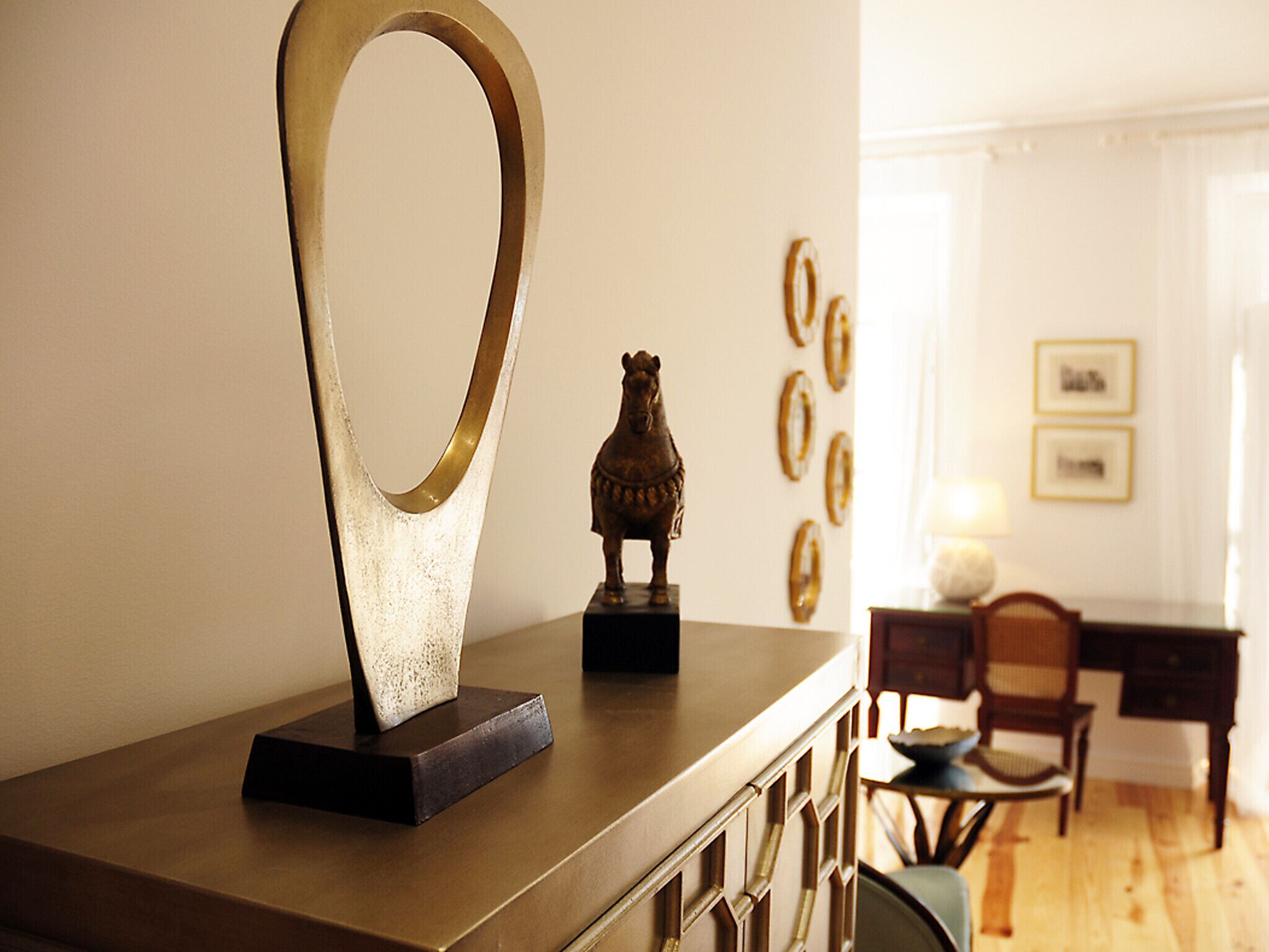 A room with a wooden dresser, a decorative metal piece, and a small statue. There are framed pictures on the wall and a dining area in the background.