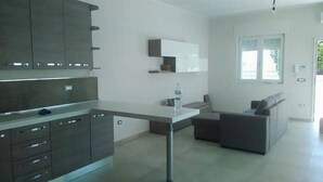 Cocina moderna con armarios oscuros, encimera blanca y horno empotrado. Cuenta con una pequeña zona de comedor con un sofá gris y una ventana que proporciona luz natural.