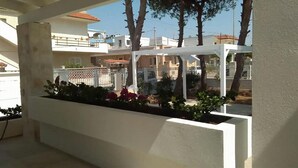 Un espacio exterior cubierto con una pérgola blanca, una jardinera con flores y vistas a edificios residenciales al fondo.