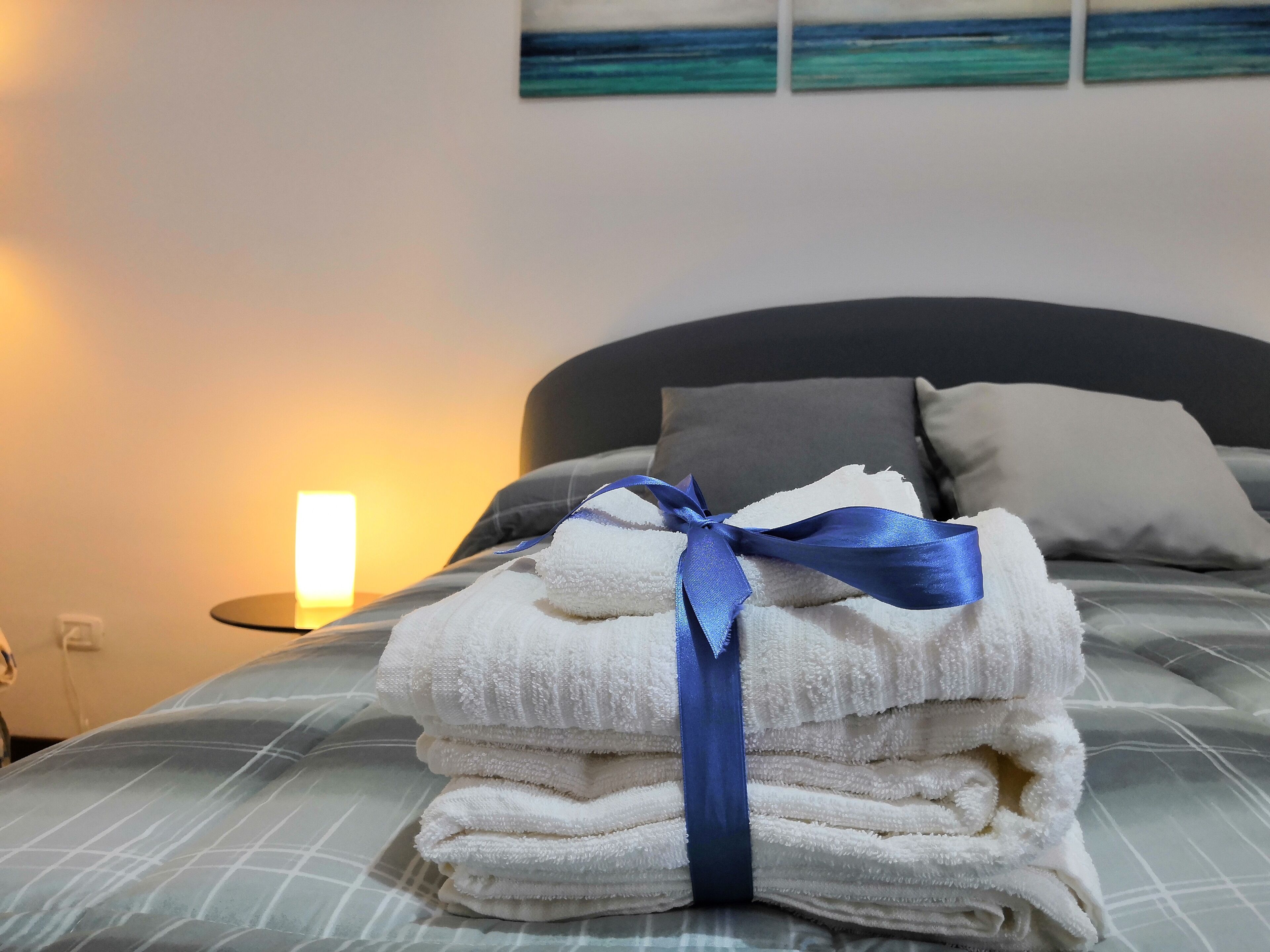 A neatly made bed with a stack of white towels tied with a blue ribbon, a gray headboard, and a bedside lamp.