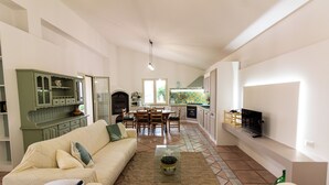 A spacious living area with a sofa, coffee table, and a television. A kitchen with a tiled backsplash and a dining area with chairs and a table are visible in the background. - Ninfa Charming Accomodation in the Eastern Coast (Floridia)