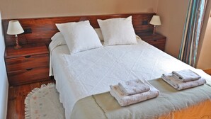 Ein ordentlich bezogenes Bett mit weißen Leinen, zwei Nachttischlampen und ein kleiner Holfnachtschrank mit einer Lampe.