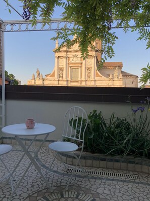 Um pátio com uma mesa e cadeira de metal branco, um vaso cor-de-rosa e vista para um edifício histórico com estátuas.