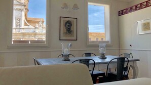 A dining area with a table and chairs, two windows with views of buildings, and a framed portrait on the wall.
