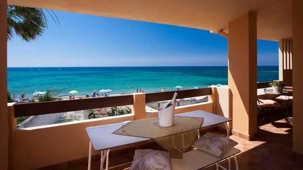 Ein Balkon mit Tisch und Stühlen mit Blick auf einen Strand und klares blaues Wasser sowie entfernte Segelboote.