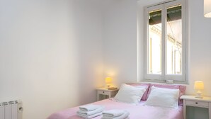Ein ordentlich bezogenes Bett mit rosa-weißer Bettwäsche, zwei Nachttischlampen und ein Fenster mit Jalousien.