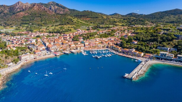 Vista aérea de un pueblo costero con un puerto deportivo, edificios y montañas al fondo.