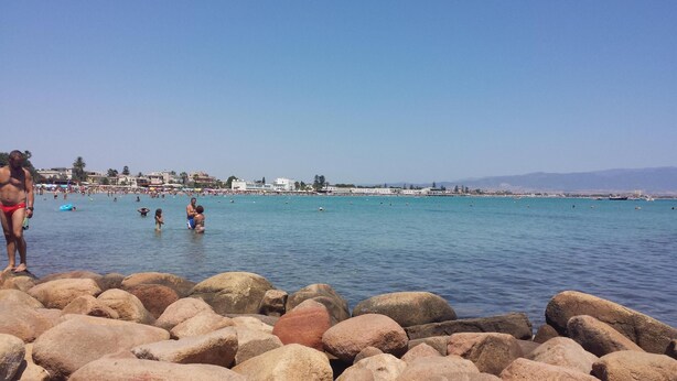 Ein Strand mit großen Felsen im Vordergrund und einer Menschenmenge im Wasser.