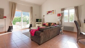 A spacious living room with a large sofa, a flat-screen TV, and a dining area visible through a sliding glass door. - Delightful villa for 4 people at Domaine Les Rives de l'Ardèche (Vallon-Pont-d'Arc)