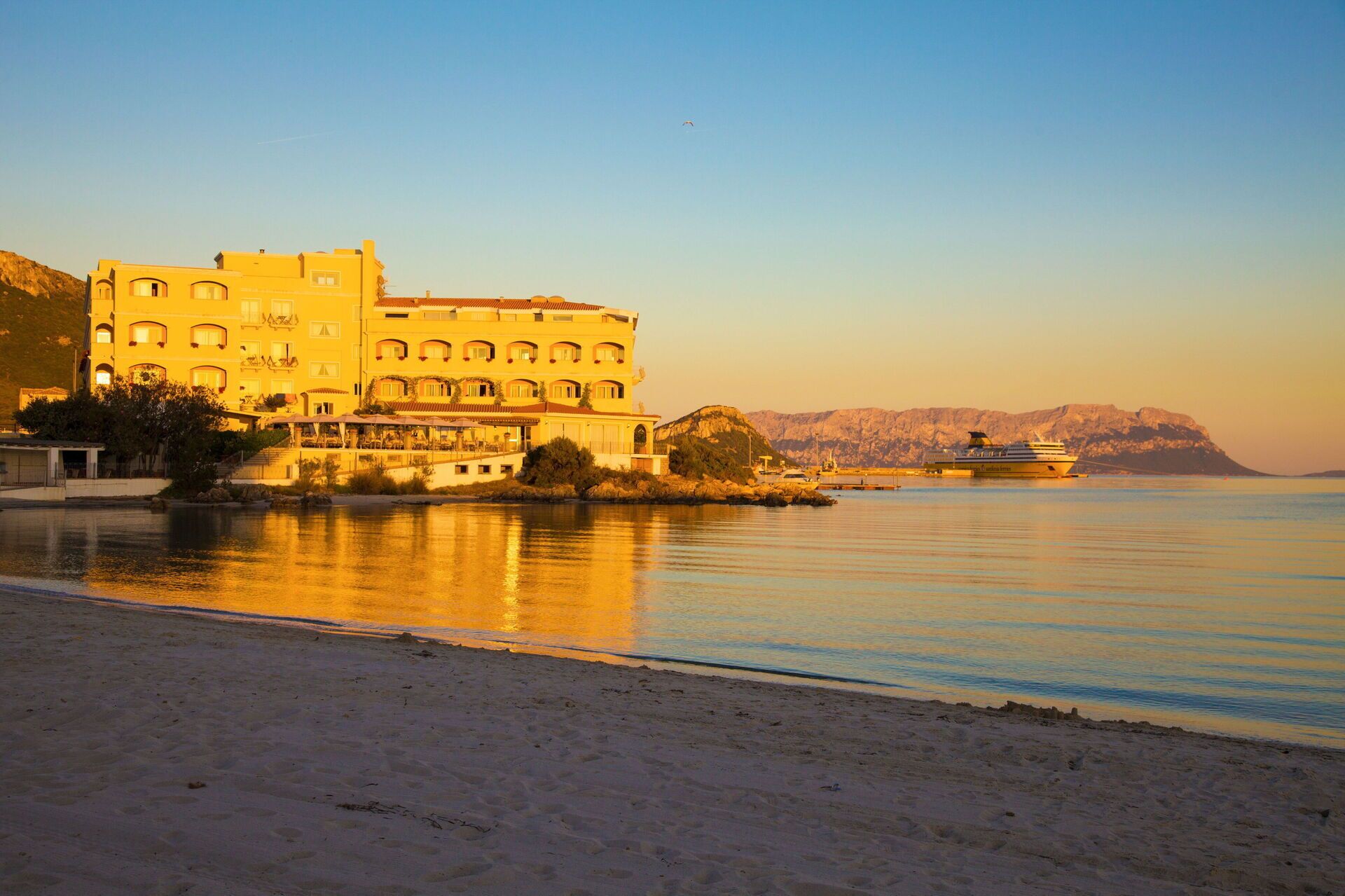 A coastal hotel with multiple balconies, a beach in the foreground, and a boat docked in the background.