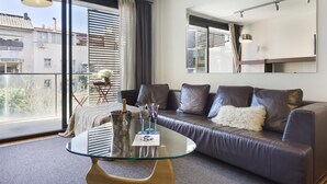A modern living room with a glass coffee table, leather sofa, and large windows offering a view of buildings and trees.