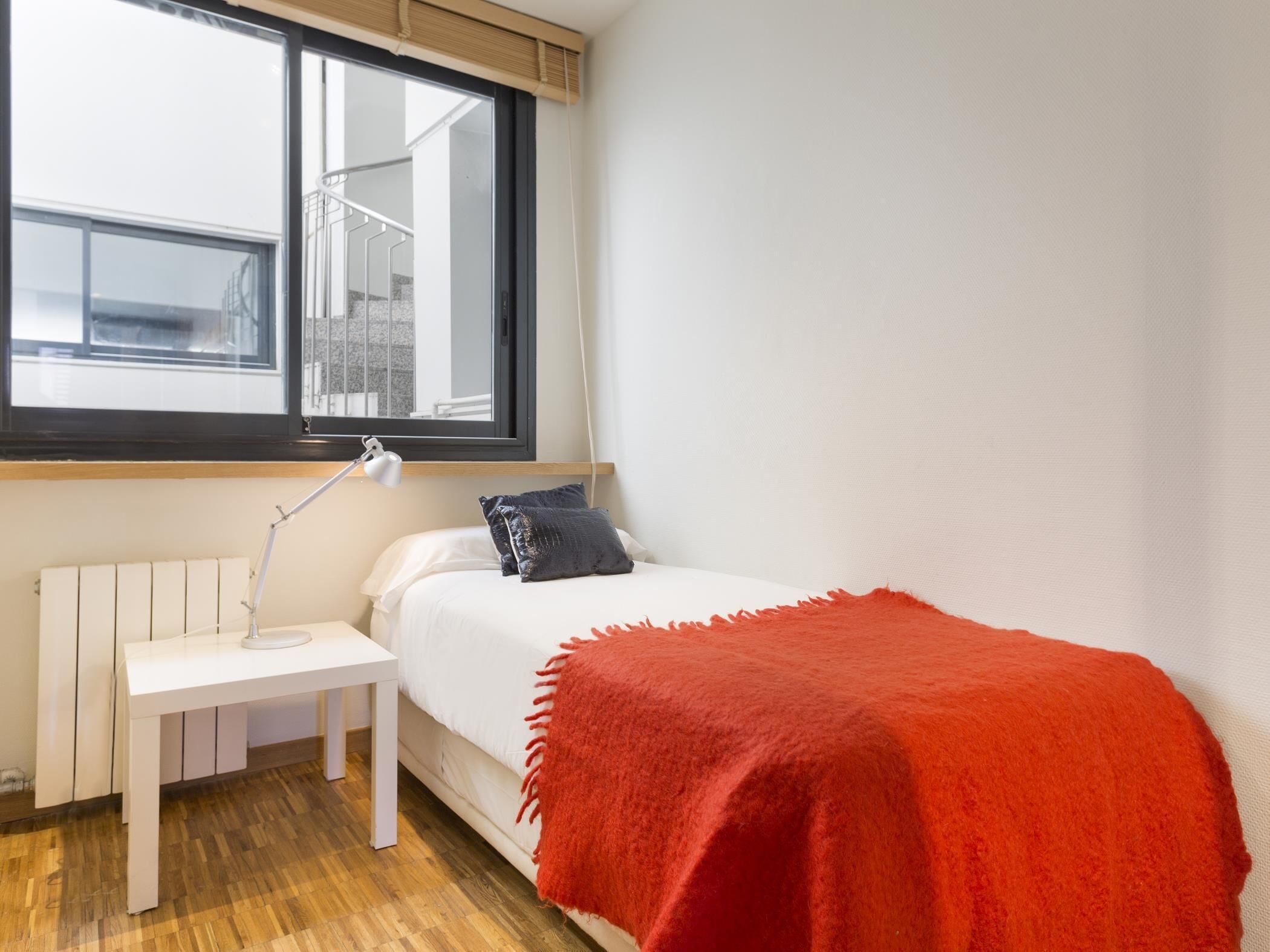 A bedroom with a bed, a bedside table, a lamp, a window, and a view of a building outside.