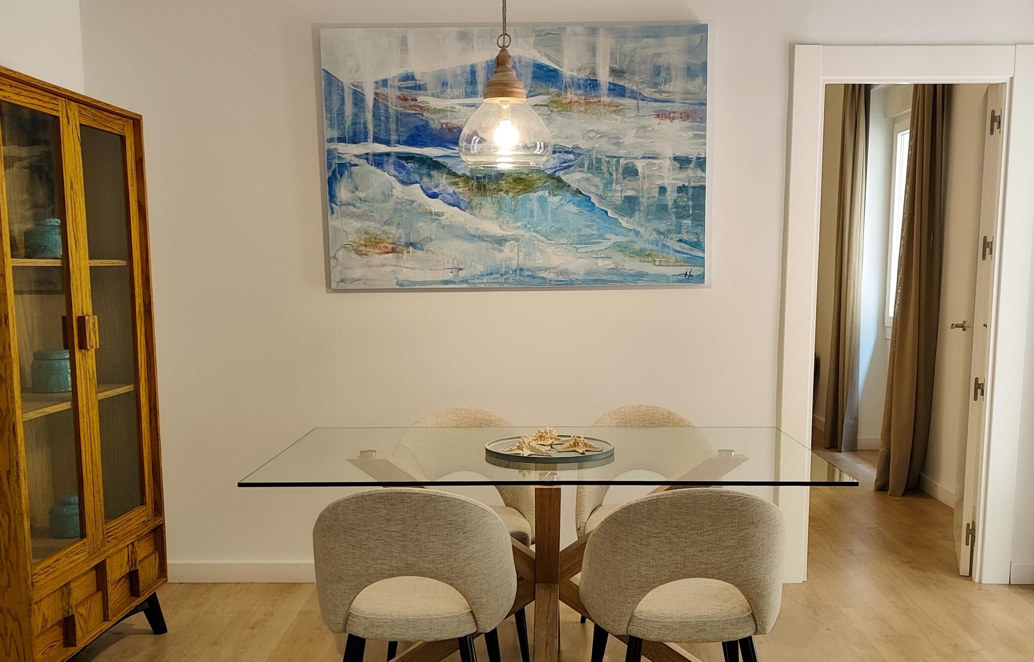 Un coin repas comprenant une table en verre, quatre chaises grises, un buffet en bois et un tableau représentant un paysage enneigé.