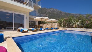 A pool area with lounge chairs and umbrellas, a stone wall, and a mountain in the background. - Elmas Apt - Luxury 3BR with Large Private Pool & Garden. 10 min Walk to Town! (Kalkan)