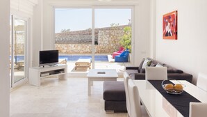A modern living room with a flat-screen TV, a coffee table, and a sofa. There is a stone wall and a view of a pool outside. - Elmas Apt - Luxury 3BR with Large Private Pool & Garden. 10 min Walk to Town! (Kalkan)