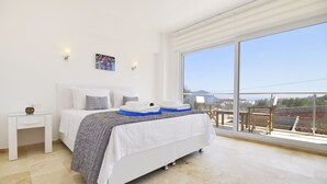 A bedroom with a large bed, a small bedside table, a bench, and a view of the outdoors. - Elmas Apt - Luxury 3BR with Large Private Pool & Garden. 10 min Walk to Town! (Kalkan)