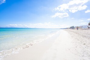 A beach with clear turquoise water, white sand, and a few buildings in the background.