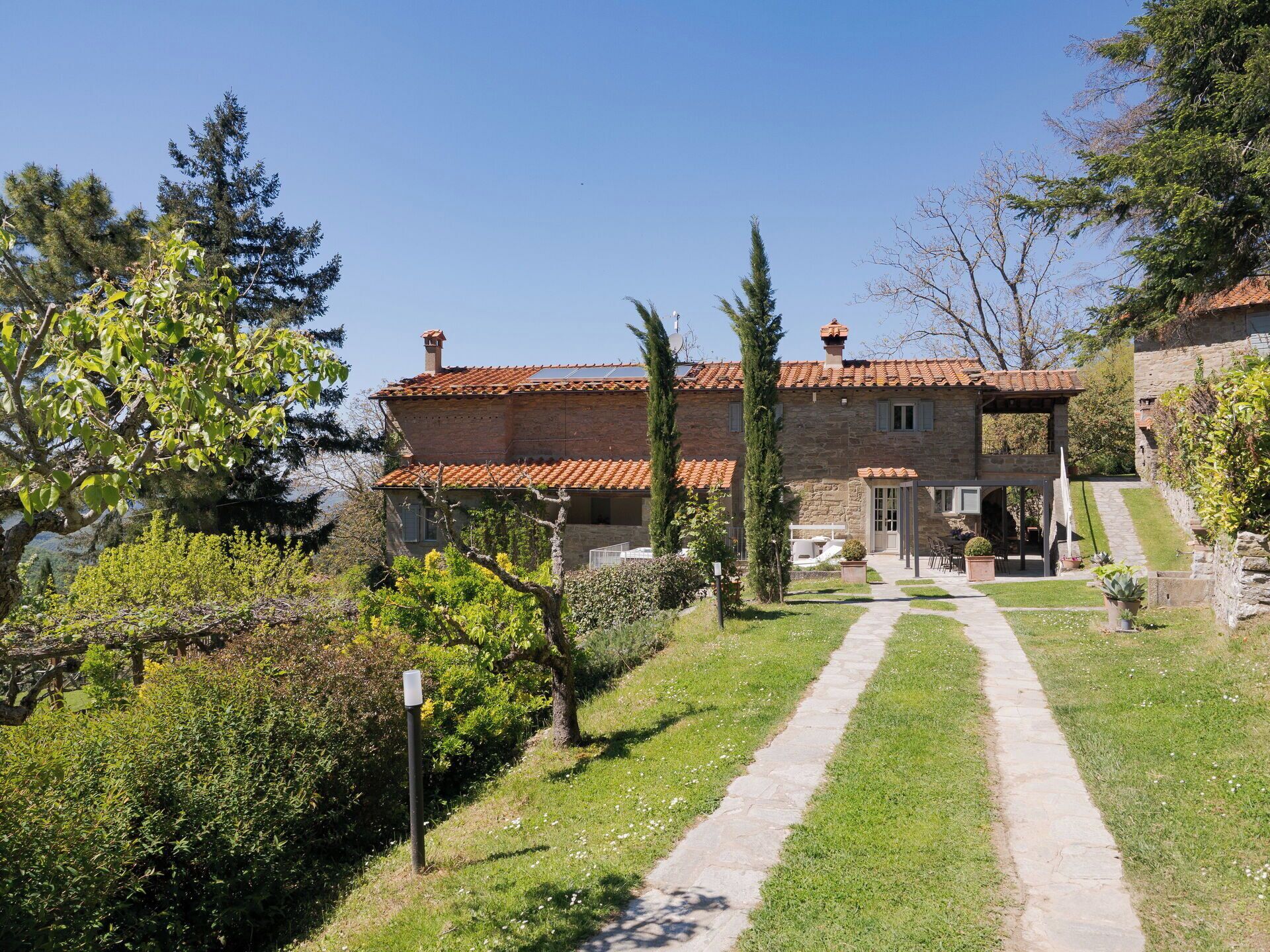 Un bâtiment en pierre avec un toit recouvert de tuiles, un chemin pavé menant à l’entrée et une variété de plantes, notamment des arbres et des arbustes.