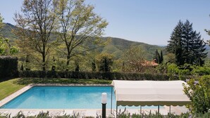 Ein rechteckiger Pool mit weißem Sonnensegel, umgeben von üppiger Vegetation und Hügeln im Hintergrund.
