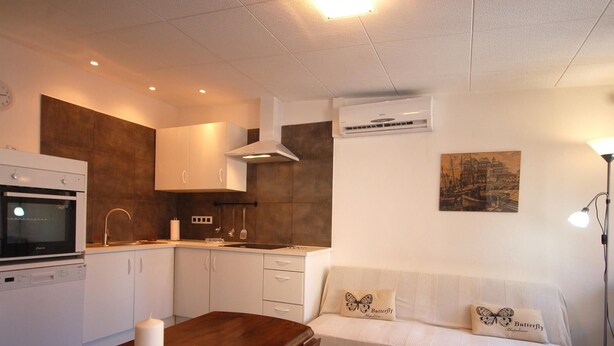 A compact kitchen with a built-in oven, microwave, and dishwasher. There's a wooden dining table with a candle and stones. A white sofa with butterfly pillows is against the wall, and an air conditioner is mounted above it. - COZY APARTMENT 70 m. from THE BEACH in TOSSA (Tossa de Mar)