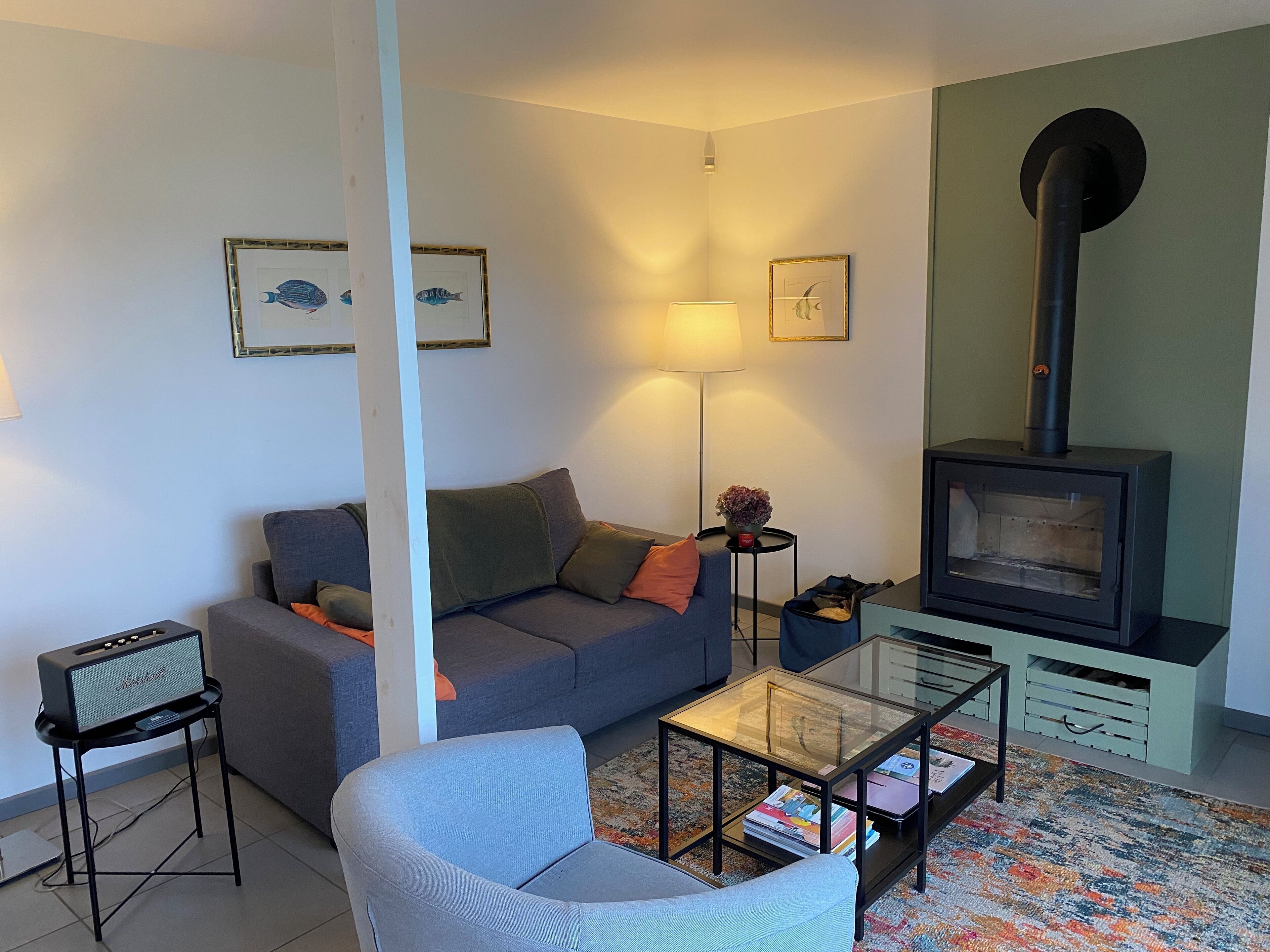 Een woonkamer met een bank, een glazen salontafel, een klein bijzettafeltje, een lamp, een speaker en een houtkachel.
