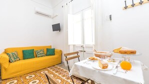 Ein helles Zimmer mit einem gelben Sofa, einem Tisch mit Speisen und Getränken sowie einem Fenster mit Vorhängen.