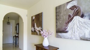 Un couloir avec un vase blanc posé sur une table en bois, deux tableaux accrochés au mur et une porte en arc donnant sur une autre pièce.