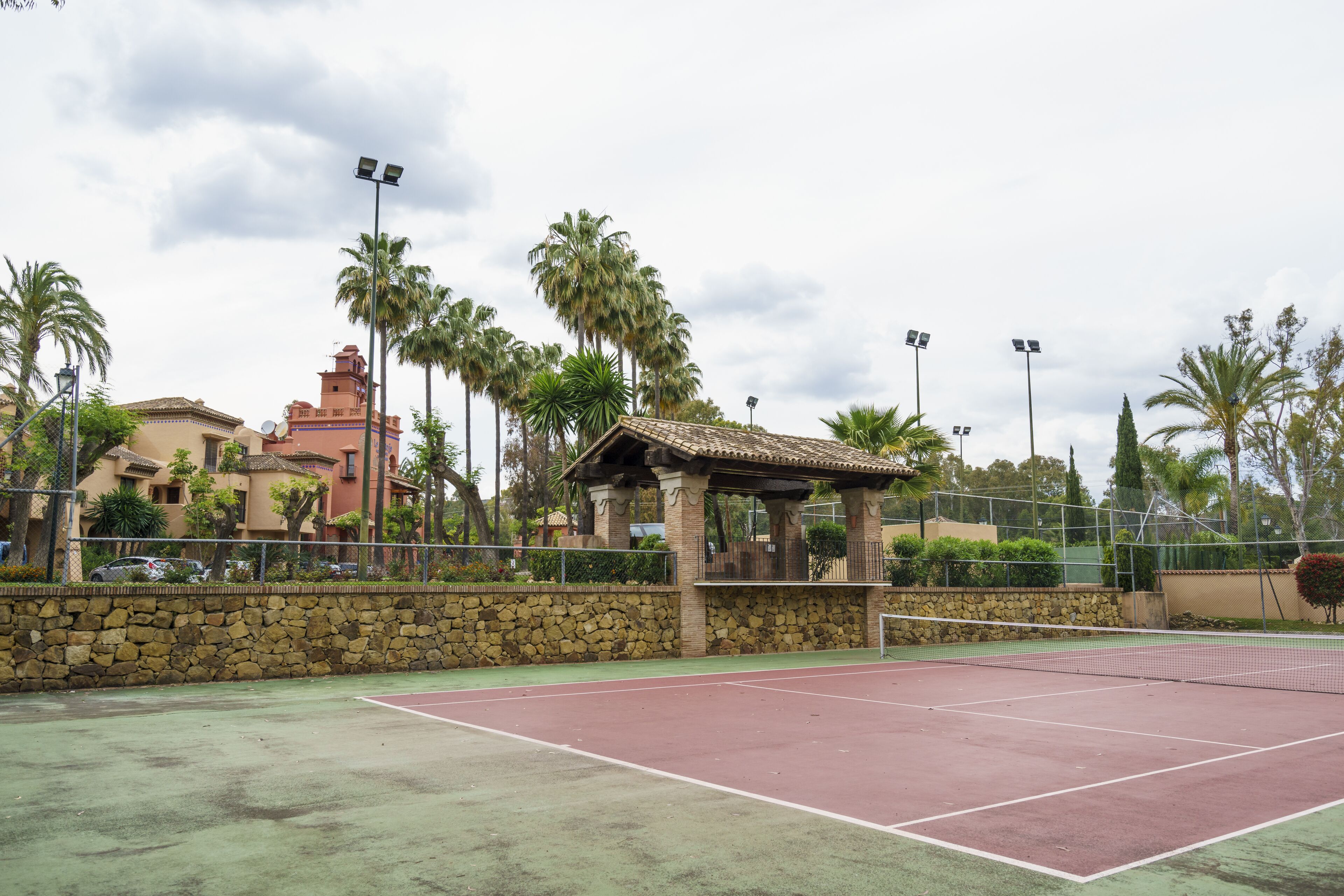 Een tennisbaan buiten met een stenen muur, palmbomen en een klein paviljoen.