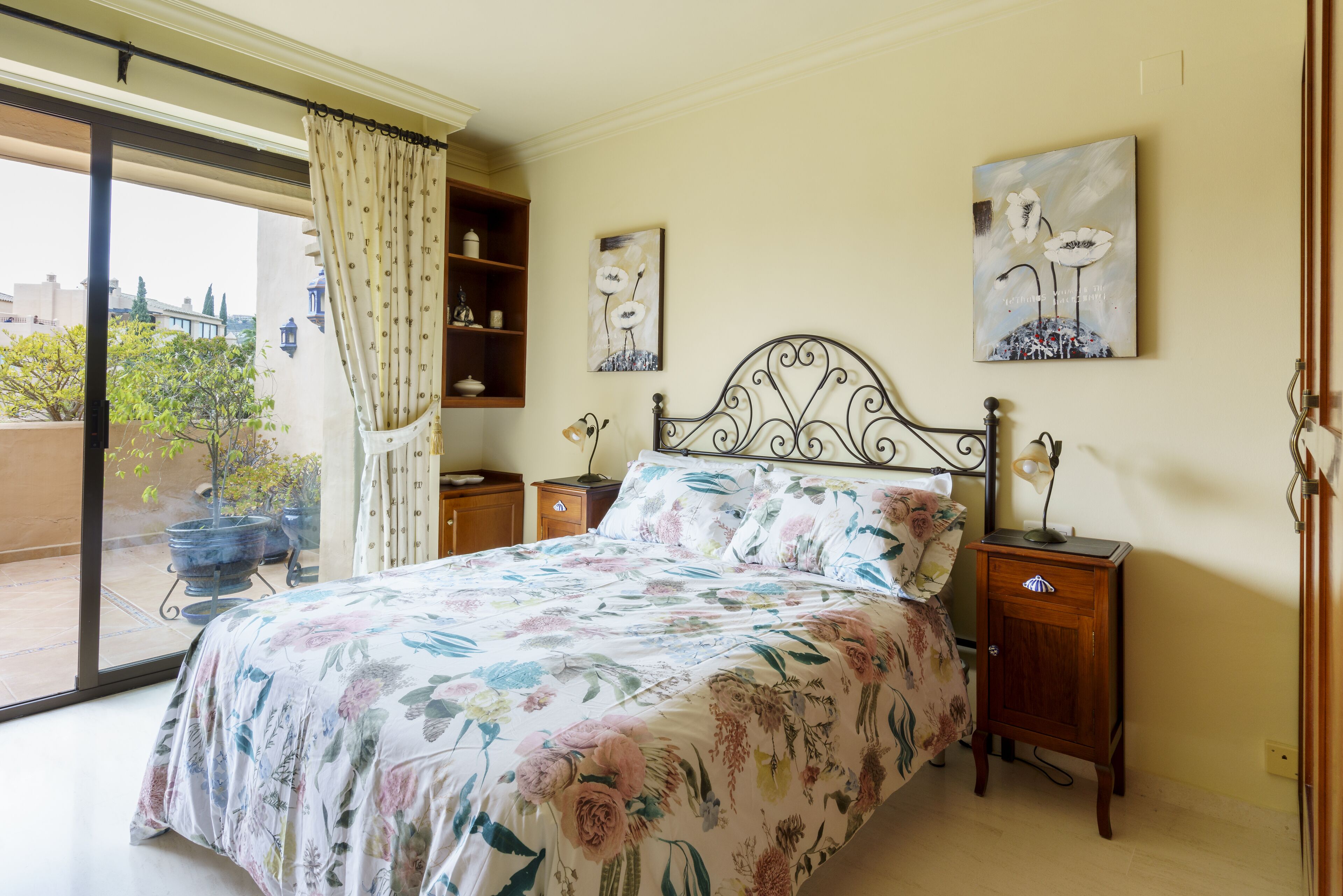 A bedroom with a bed, a nightstand, a bedside table, a window with curtains, and a view of the outside.
