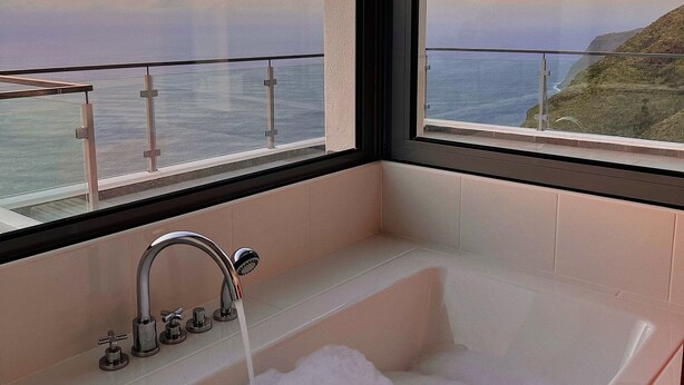 A bathroom with a bathtub filled with bubbles, a modern faucet, and a view of the ocean through large windows. - Quinta Pereira ... beautiful panoramic views and ocean sunsets  (Faja Da Ovelha)