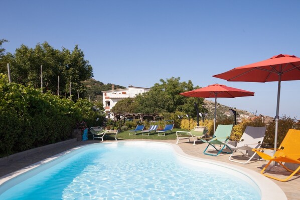A swimming pool with lounge chairs and umbrellas, surrounded by greenery and a hillside in the background.