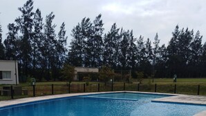 Una piscina rectangular con borde de baldosas, rodeada por una valla y árboles.
