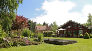 Ein gepflegter Garten mit einer Steinbank, ein Backsteinhaus mit großen Fenstern und einem strahlend blauen Himmel.