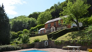 A swimming pool with a wooden deck, a stone wall, and a house with a red roof and white trim in the background. - Riversdale Lodge is set in the heart of Symonds Yat overlooking the River Wye.  (Symonds Yat)
