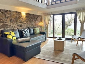A modern living room with a stone wall, a black leather sofa with patterned cushions, a wooden coffee table, and a view of the outdoors through large windows.
