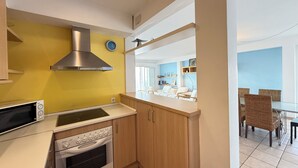Een compacte keuken met een magnetron, oven en een roestvrijstalen afzuigkap. Op de achtergrond is een eethoek met rieten stoelen en een tafel te zien.
