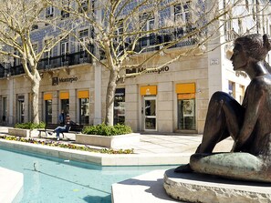 A bronze sculpture of a seated figure in an outdoor plaza with a pool, surrounded by buildings with storefronts and balconies. - Fully equipped air-conditioned apartment in central Setúbal (Setúbal)