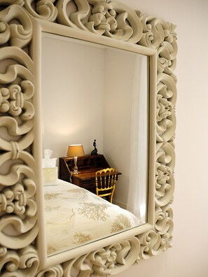 A mirror with an ornate frame reflecting a room with a bed, a lamp, and a chair.