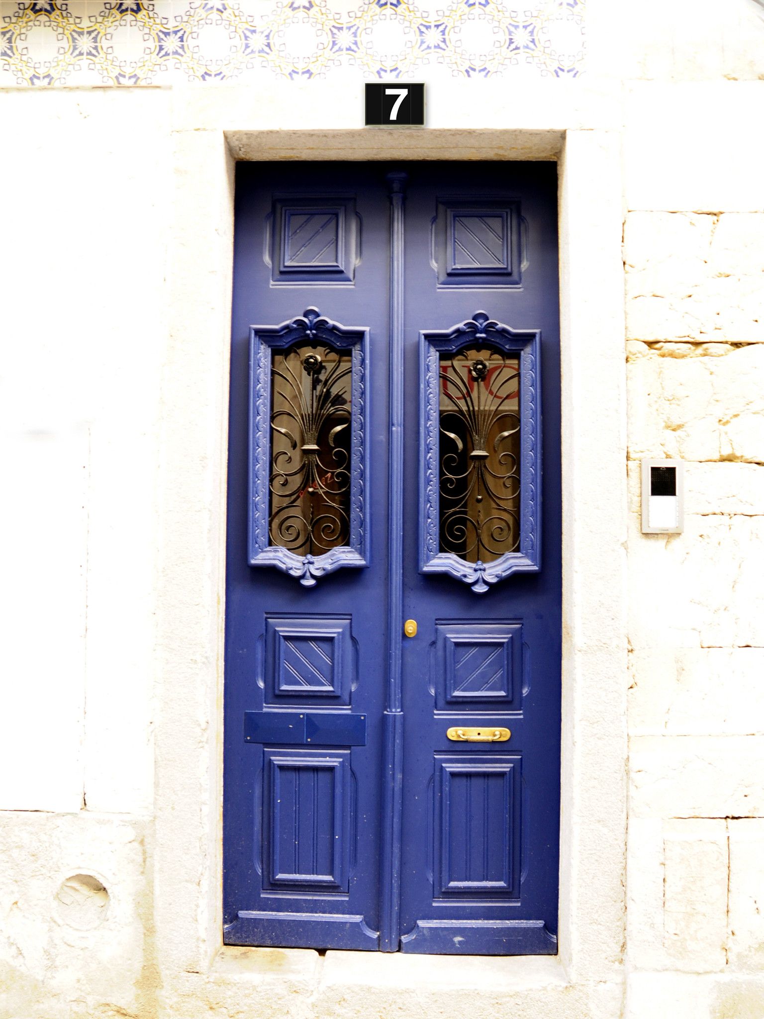 Une porte bleue à deux battants, ornée de motifs métalliques, avec le chiffre 7 au-dessus, et une petite fenêtre sur le côté droit.